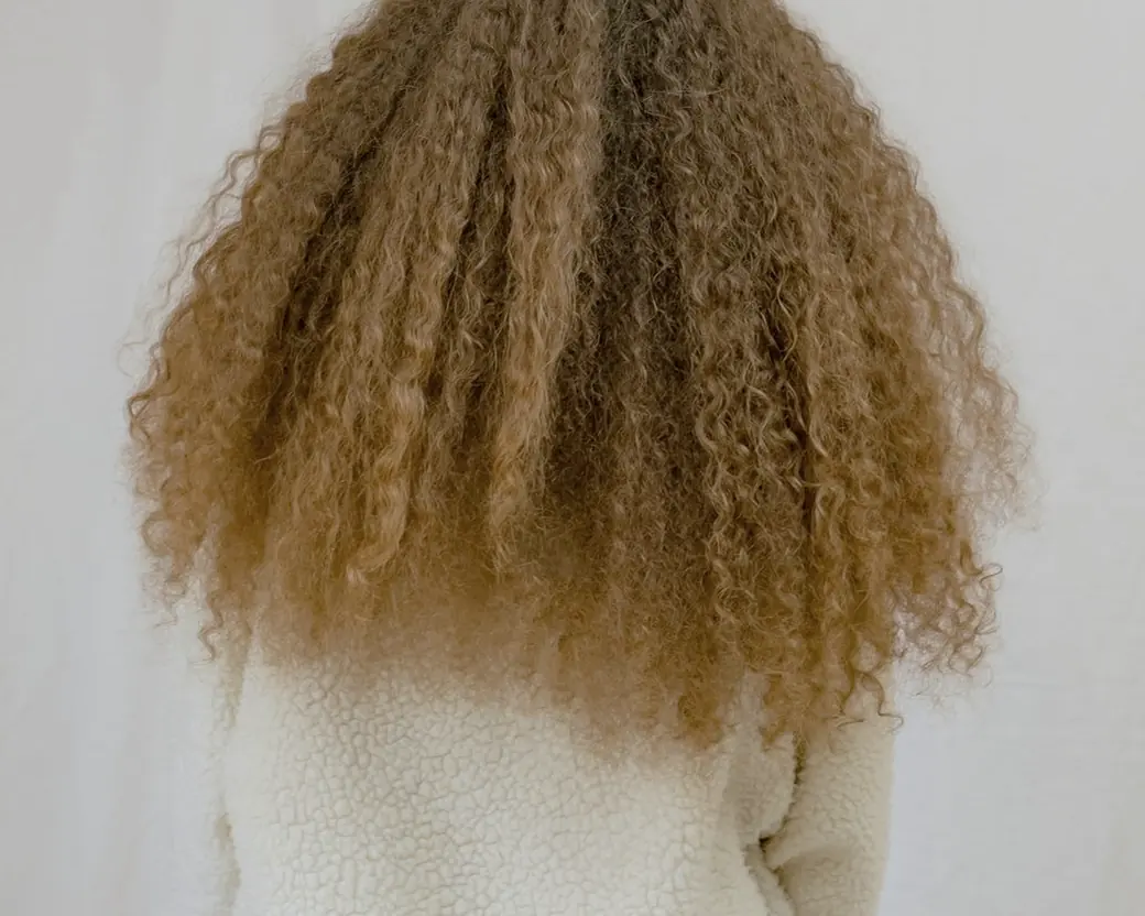 Golden-brown curly hair seen from behind, falling to the shoulders of a cream knit top, against a plain cloth backdrop.