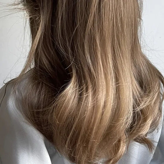 The lower length of straight brown hair with sun-lightened ends curling under onto a pale gray shoulder, against a plain wall.